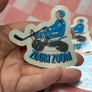 Könnte beinhalten: Ein holografischer Aufkleber mit einem Hockeyspieler auf einem vierrädrigen Fahrzeug. Der Spieler trägt eine blaue Uniform mit der Nummer 71. Der Aufkleber hat die Worte "ZOOM ZOOM" in Blau, mit einem Hockeyschläger und einem Puck.