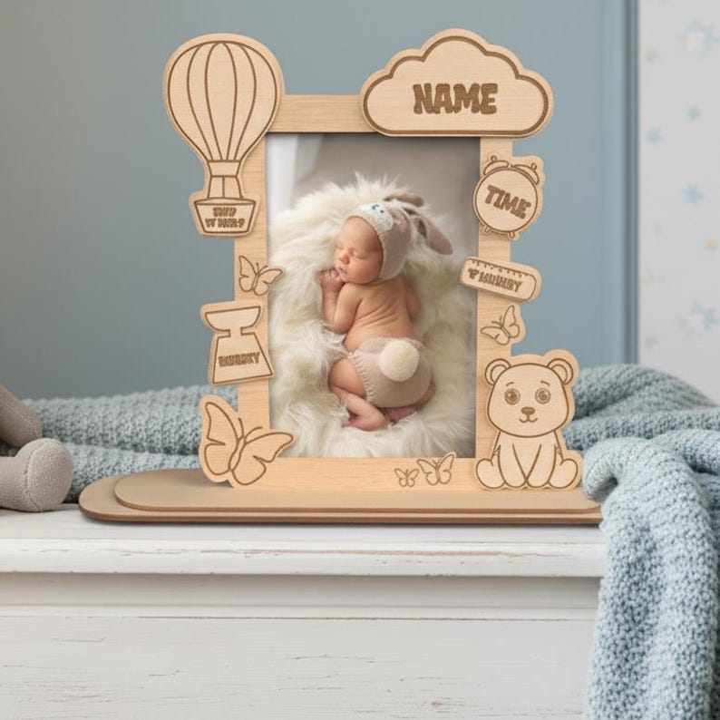 May include: Wooden baby photo frame with a hot air balloon, cloud with "NAME" text, butterflies, a bear, and an alarm clock. The frame holds a photo of a sleeping baby. The frame is made of light-colored wood.