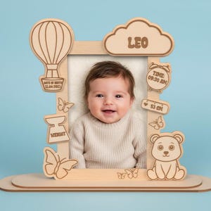 Puede incluir: Marco de fotos de bebé de madera con detalles cortados con láser. Presenta un globo aerostático, una nube con "LEO", un reloj y un oso. Incluye la fecha, la hora, el peso y una medida de 53 cm. El marco contiene un bebé sonriente.