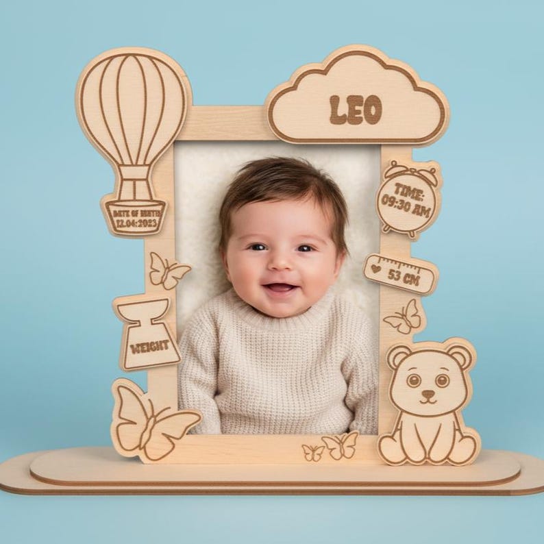May include: Wooden baby photo frame with laser-cut details. Features a hot air balloon, cloud with "LEO", a clock, and a bear. Includes the date, time, weight, and 53 cm measurement. The frame holds a smiling baby.