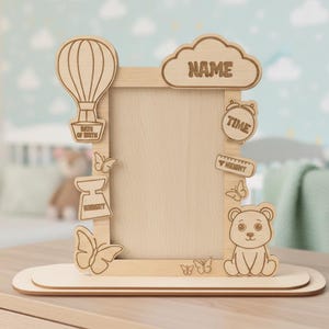 May include: Wooden baby photo frame with decorative cutouts of a hot air balloon, butterflies, a bear, and a cloud with the word "NAME". Includes cutouts for "DATE OF BIRTH", "WEIGHT", "TIME", and "HEIGHT".