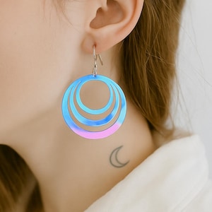 May include: Blue, pink, and purple gradient hoop earrings. The earrings have three concentric circles, each decreasing in size. The earrings are hanging from a silver hook. The earrings are worn on the ear.