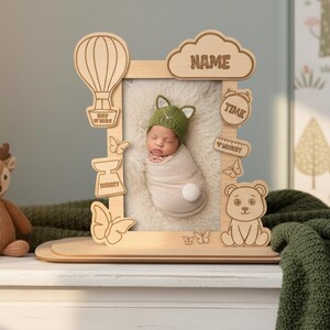 May include: A wooden baby photo frame with laser-cut details, including a hot air balloon, butterflies, and a bear. The frame holds a photo of a newborn baby wearing a green hat. The word "NAME" is on the top of the frame.