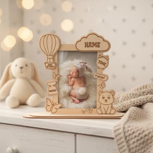May include: Wooden baby photo frame with a hot air balloon, cloud, bear, and butterfly cutouts. The frame holds a photo of a newborn baby. The frame has the word "NAME" on the cloud cutout and other details.
