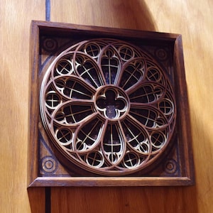 May include: A wooden, layered wall art piece featuring a circular, gothic rose window design. The artwork is framed in a dark brown square, with a lighter wood colour used for the window's intricate details. The piece is mounted on a wooden wall.