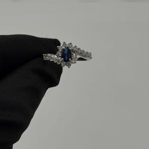 May include: A silver ring featuring a dark blue oval gemstone surrounded by smaller clear stones. The ring's design includes a halo of clear stones around the central gem and additional stones along the band, creating a sparkling effect. Jewelry.