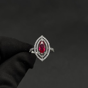 May include: A silver ring featuring a marquise-cut, deep red gemstone, surrounded by a double halo of small, clear gemstones. The ring's design is symmetrical and elegant, set against a dark background.