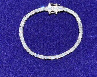 Op de afbeelding: Een zilveren armband met rechthoekige, heldere edelstenen, in een rij gezet. De armband is gesloten en rust op een diepblauwe fluwelen ondergrond. Het woord "LAVISH" staat bovenaan de afbeelding.