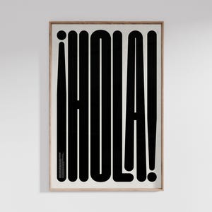 Op de afbeelding: Een ingelijste kunstprint met het woord "HOLA!" in vette, zwarte, gestileerde letters. Het uitroepteken en de letter "O" zijn cirkelvormig. De print zit in een licht houten lijst en hangt aan een witte muur.