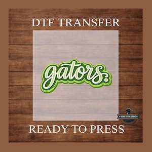 May include: A DTF transfer with the word "gators" in a layered green and white design. The text "DTF TRANSFER" is above the design, and "READY TO PRESS" is below. The background is a wood grain pattern.