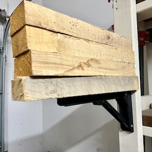May include: A set of five wooden planks are supported by a black metal folding shelf bracket. The bracket is attached to a white wall.