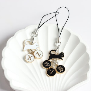 May include: Two cat-themed phone charms. One charm features a white cat with a silver-tone clasp and a black cord. The other charm has a black cat. Both charms have lettered pendants with gold rims. The charms are displayed on a white, scalloped dish.