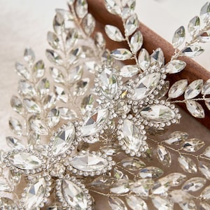 Puede incluir: Un tocado de novia plateado adornado con cristales transparentes y brillantes. El tocado presenta un diseño floral y de hojas, con grandes cristales de talla marquesa en el centro y cristales más pequeños que delinean las hojas.