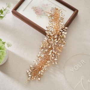 May include: A gold-toned hair accessory with clear crystal accents, shaped like a floral vine. The piece is displayed on a white surface near a framed picture and a decorative clear acrylic circle.