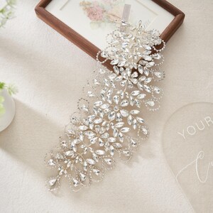 May include: A silver-toned bridal headpiece with clear, sparkling crystals. The headpiece features a floral and leaf design, with a cascade of crystals. The piece is designed to be worn on the side of the head, adding a touch of elegance.