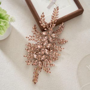 Puede incluir: Un peine para el cabello de novia en oro rosa con un diseño floral. El peine presenta grupos de cristales brillantes y delicados acentos de hojas. Los cristales son de color rosa suave y el metal tiene un tono dorado cálido. El peine está diseñado para ocasiones formales.