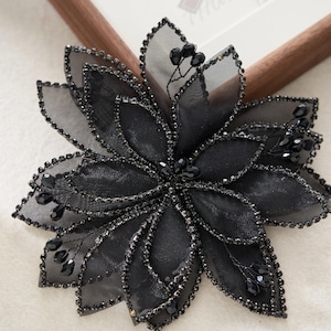 May include: A black floral embellishment with layered petals and beaded edges. The center and edges are adorned with small, dark beads. The flower is made of a sheer fabric, creating a delicate and elegant appearance. It is likely used for crafting or fashion.