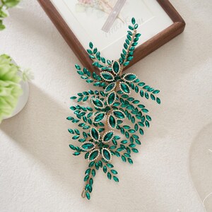 May include: A decorative hair accessory featuring emerald green gemstones arranged in floral and leaf patterns. The piece is accented with small, clear crystals and gold-toned metal, creating a sparkling, elegant design. Ideal for weddings or special occasions.