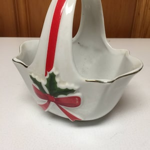 May include: White ceramic basket with a red stripe on the handle and a holly and bow design. The basket has a scalloped edge with gold trim, suitable for festive decor or as a decorative item.