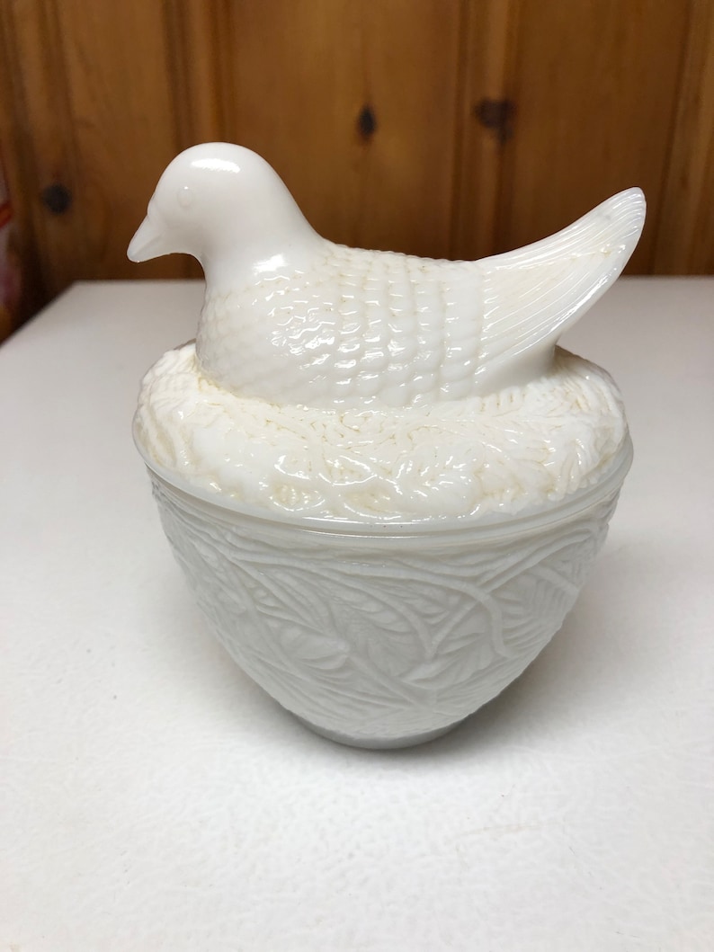 May include: A white, vintage-style bird-shaped trinket box. The bird sits atop a nest-like base, with detailed feather and leaf patterns. The lid is a bird, and the base is a bowl. The overall design is a classic, decorative piece.