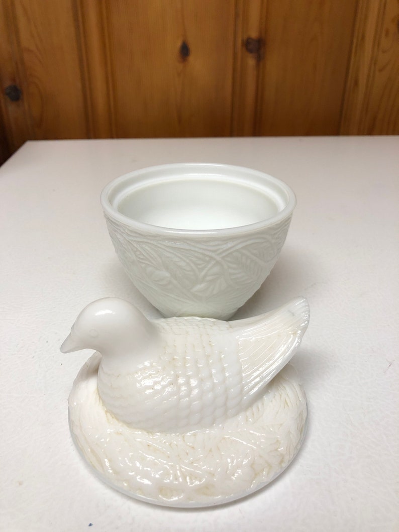 May include: A white ceramic dove-shaped trinket box with a matching lid. The lid is a dove sitting on a nest. The box has a textured, embossed design. The box is open, revealing the interior.