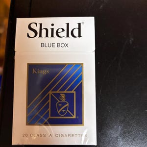 May include: A white and blue Shield Blue Box of Kings cigarettes. The box features a blue and gold design with the text "Shield" at the top. The box contains 20 Class A cigarettes.
