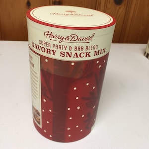 May include: A cylindrical container of Harry & David Savory Snack Mix. The container has a red background with white snowflake and floral designs. The top and bottom rims are white with a red border. The text "Harry & David" and "Super Party & Bar Blend Savory Snack Mix" are printed on the container.