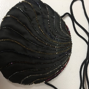 Puede incluir: Un bolso de noche negro de cuentas con cierre de cordón negro y una correa de cordón negro. El bolso está decorado con cuentas doradas, plateadas e iridiscentes en forma de onda.