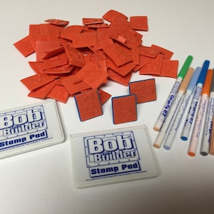 May include: A set of Bob the Builder themed toys, including a stack of orange rubber stamps, two white stamp pads with the text "Bob the Builder Stamp Pad", and a set of ten colorful markers.