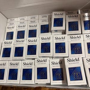 Puede incluir: Una colección de cajetillas de cigarrillos rectangulares blancas etiquetadas como Shield Blue Kings Box, con un gráfico cuadrado azul con líneas diagonales doradas y un emblema de escudo. Cada cajetilla indica 20 cigarrillos de clase A.