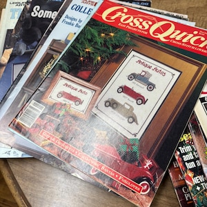 May include: A stack of vintage cross-stitch pattern books. The top book features a red cover with the words "Cross Quick" and "Antique Autos" designs. Other books have titles like "Some" and "Trim and fun".