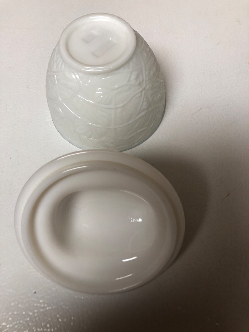 May include: A white, vintage-style bowl and lid set. The bowl has a textured leaf pattern, and the lid is oval-shaped with a handle. The set is made of opaque white glass.