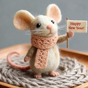 May include: A handcrafted felt mouse figurine with a peach-colored scarf, holding a small sign that reads "Happy New Year!". The mouse has large ears and a long tail. The figurine is set on a woven surface.