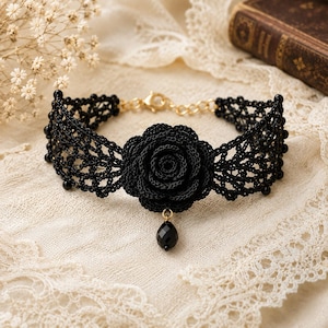 May include: A black lace choker necklace featuring a central crocheted rose motif and a dangling black teardrop bead. The band is crafted from intricate black thread in a web-like pattern, finished with a gold-toned metal chain and lobster clasp.