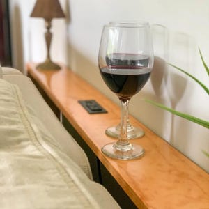 May include: Two clear wine glasses filled with red wine sit on a wooden shelf. The shelf is a light brown color and is next to a white wall. A small lamp with a brown lampshade is in the background.
