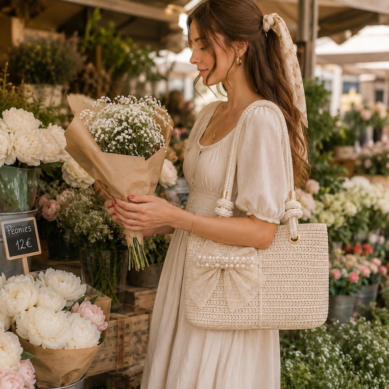 May include: A cream-colored woven tote bag featuring a decorative bow adorned with white faux pearls on the front. The bag has two sturdy braided handles attached with gold-tone hardware, a structured rectangular shape, and a textured straw-like finish.