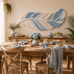 Pode incluir: Uma grande tape&ccedil;aria horizontal em forma de pena, feita com fio creme e azul claro num padr&atilde;o de croch&eacute; texturizado. A pe&ccedil;a est&aacute; montada sobre uma mesa de jantar de madeira decorada com vasos de cer&acirc;mica e roupa de mesa.