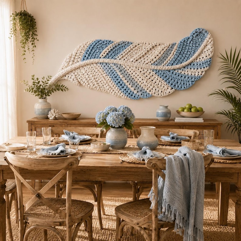 Pode incluir: Uma grande tape&ccedil;aria horizontal em forma de pena, feita com fio creme e azul claro num padr&atilde;o de croch&eacute; texturizado. A pe&ccedil;a est&aacute; montada sobre uma mesa de jantar de madeira decorada com vasos de cer&acirc;mica e roupa de mesa.