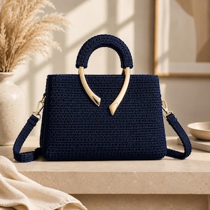 May include: A dark navy blue crochet handbag featuring a structured rectangular body and a matching crocheted top handle. The handle is accented with two polished gold-tone metal hardware pieces that curve downward. It includes a detachable shoulder strap.
