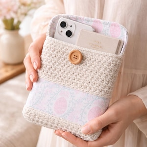 Op de afbeelding: Een crèmekleurige gehaakte telefoonhoes met een stoffen paneel met bloemenpatroon. De hoes is vastgezet met een houten knoop. Een smartphone en een klein notitieboekje zijn zichtbaar in de hoes. Op het notitieboekje staat het woord "JOURNAL".