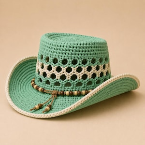 May include: A mint green crocheted hat featuring a wide brim with a cream-colored edge. The crown includes a decorative band of open-weave hexagonal patterns and a string of brown and cream wooden beads tied in a knot at the front.
