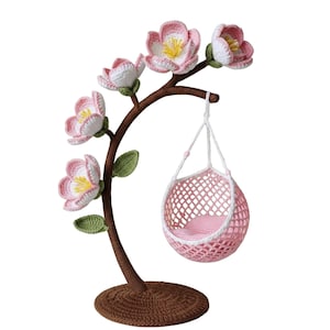 Op de afbeelding: Een handgemaakt decoratief haakwerk met een bruine tak met roze en witte bloemen en groene bladeren. Een kleine, roze haakschommel hangt aan de tak. De basis is een bruin, cirkelvormig geweven ontwerp.