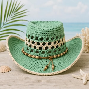 Può includere: Un cappello da cowboy all'uncinetto color verde menta con tesa larga e bordo color crema. La corona presenta una fascia decorativa con motivo esagonale traforato color crema e un sottile cordino marrone con piccole perline di legno annodato sul davanti.