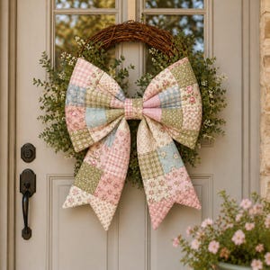 May include: A door wreath with a large, quilted bow in pastel pink, blue, and green patchwork patterns. The bow is set against a circular wreath of greenery and twigs, creating a charming, country-style accent for a door.