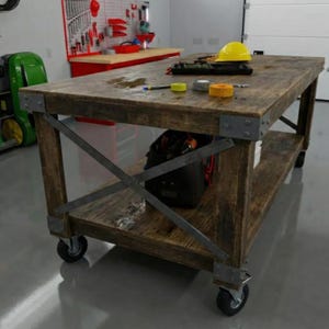 May include: A large, rustic wooden workbench with metal corner brackets and a lower shelf. The workbench has wheels and is topped with tools, tape, and a yellow hard hat. A black tool bag sits on the shelf below.
