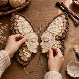 Peut inclure: Un projet au crochet compos&eacute; de deux pi&egrave;ces beiges en forme de visage avec des yeux ferm&eacute;s et des l&egrave;vres brod&eacute;s, fix&eacute;es sur de grandes ailes de papillon superpos&eacute;es dans des tons cr&egrave;me et brun fonc&eacute;, mont&eacute;es sur une fine armature m&eacute;tallique.
