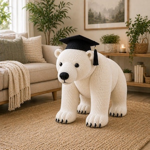 May include: A large, white crocheted polar bear wearing a black graduation cap with a tassel. The bear has black eyes, a black nose, and black claws. It stands on all fours on a woven jute rug, with a neutral-toned living room in the background.