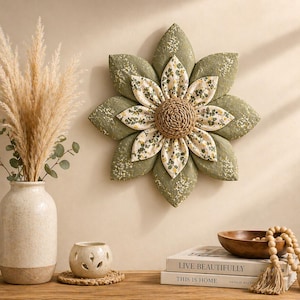 Puede incluir: Un gran adorno de pared con forma de flor de tela, con p&eacute;talos verde oliva y un centro con estampado floral. El centro es de material tejido. La flor est&aacute; montada en una pared beige.