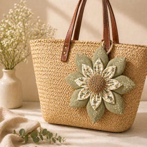 Puede incluir: Un bolso tote de paja tejida con asas de cuero marr&oacute;n y una gran flor decorativa de tela. La flor tiene p&eacute;talos verdes y blancos con un centro tejido. El bolso es de color beige natural.