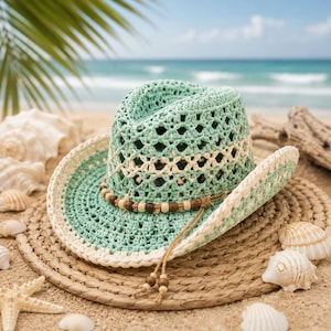 May include: A turquoise crochet cowboy hat with a cream-colored brim and accents, adorned with a wooden bead band. The hat rests on a woven straw hat, surrounded by seashells and sand, with a beach and ocean backdrop.